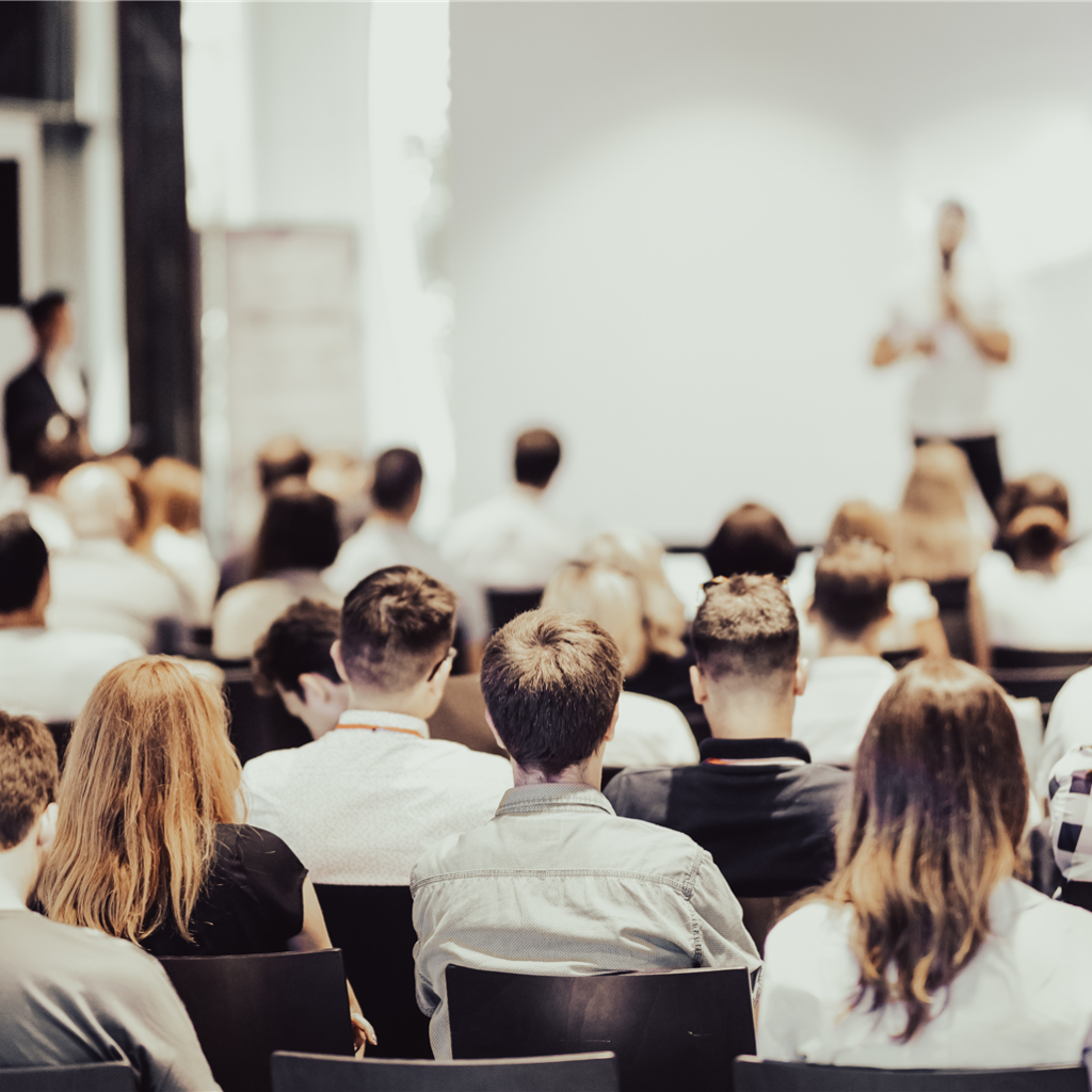 Hörsaal Publikum von hinten in einem Saal, vorne unscharfe Person stehend und sprechend.