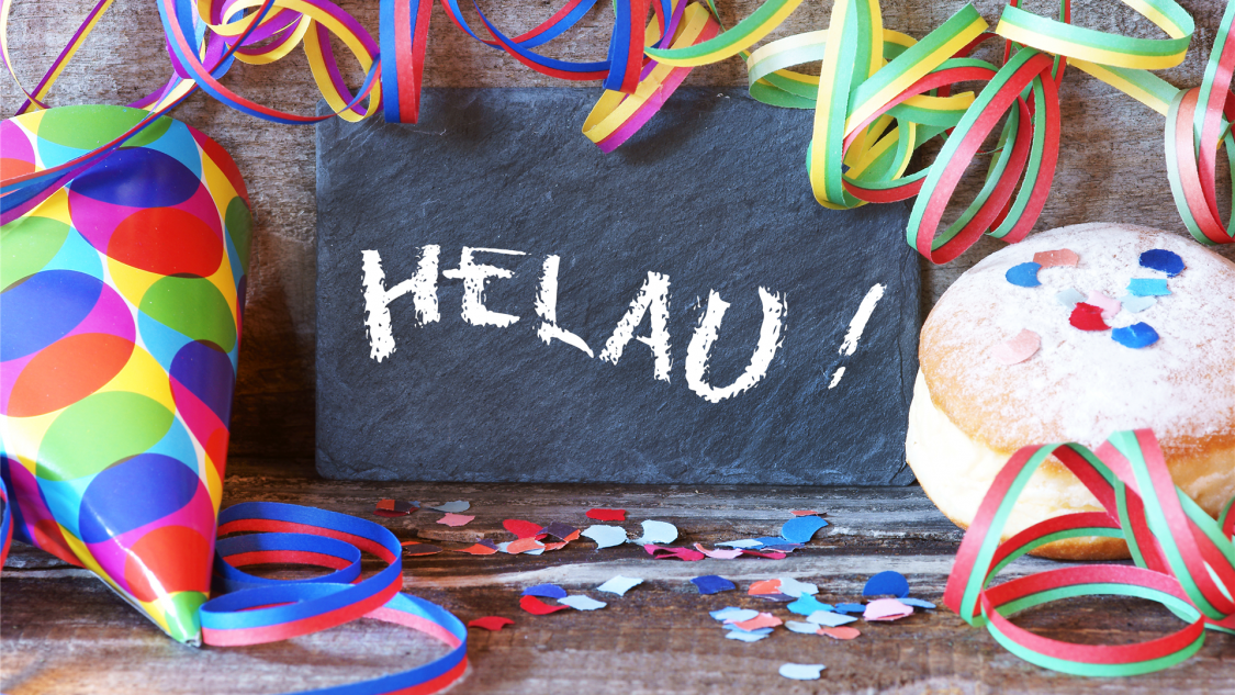 Kreidetafel mit Helau-Aufschrift. Daneben bunte Luftschlange, Kreppel und bunter Partypapphut.